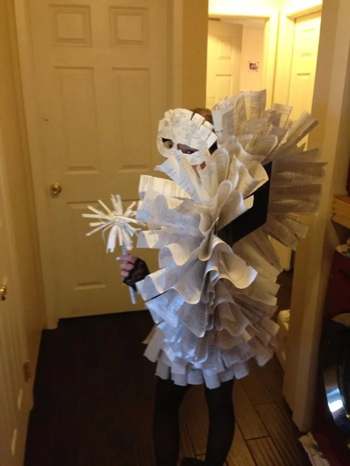 Child wearing a creative Halloween costume made from curled paper pages, holding a matching paper flower prop indoors.