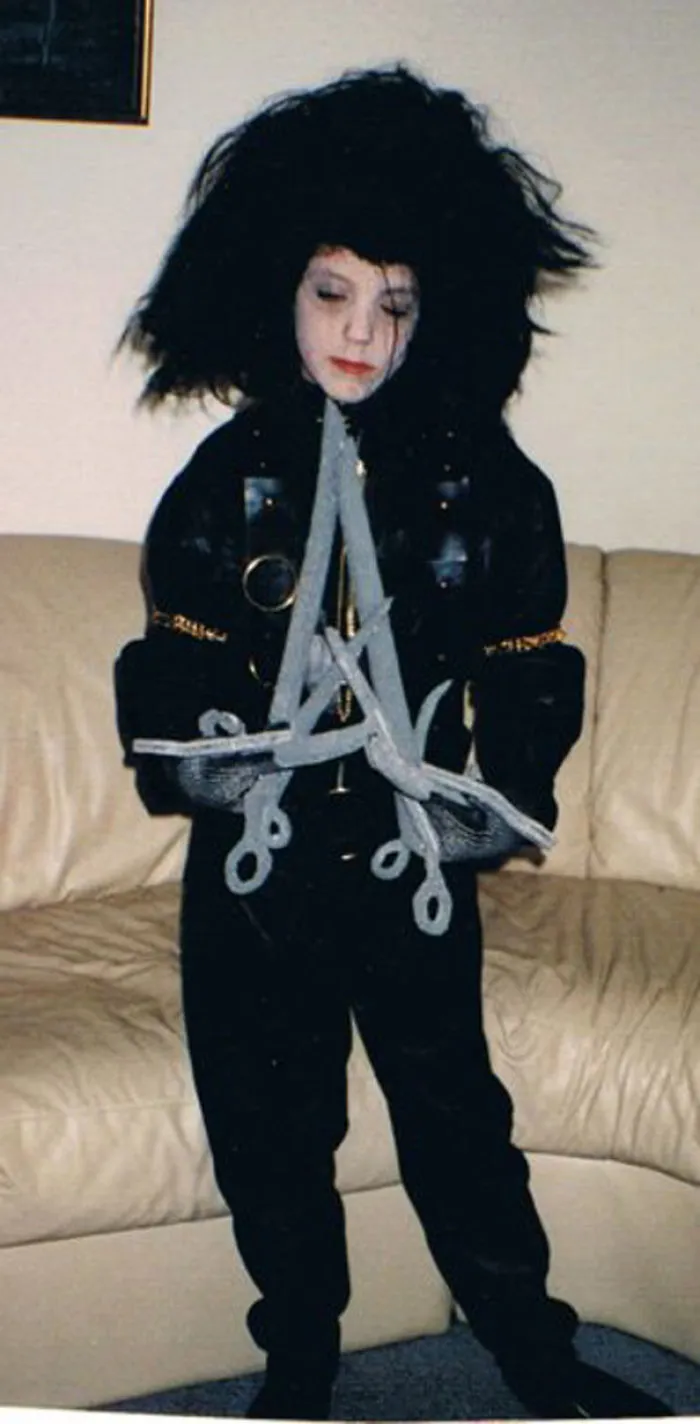 Child in an elaborate Halloween costume with black outfit, large wild wig, and gray scissor prop for children's Halloween costume ideas.