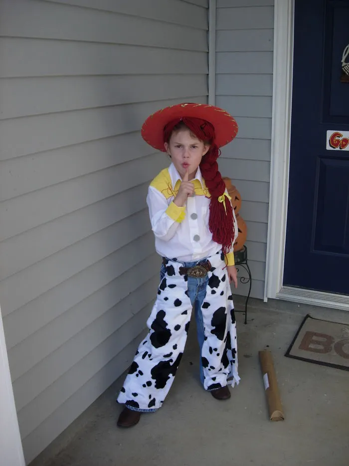 Child in a cow-print Halloween costume with a red hat and braided wig, posing for children's Halloween costume ideas.