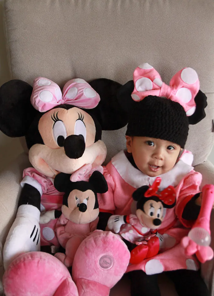 Baby dressed in Minnie Mouse costume sitting with Minnie Mouse plush toys for children's Halloween costume ideas.
