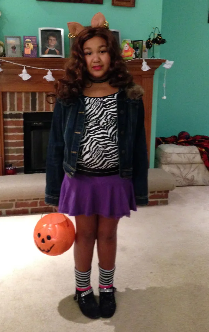 Child wearing Halloween costume with cat ears, zebra print top, purple skirt, and holding pumpkin bucket for children's Halloween costume ideas.