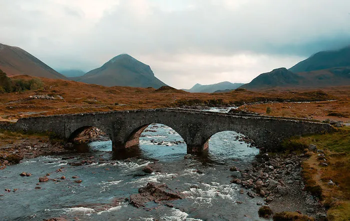 Ancient mystical stone bridge over flowing river in a remote mountainous landscape under cloudy sky.