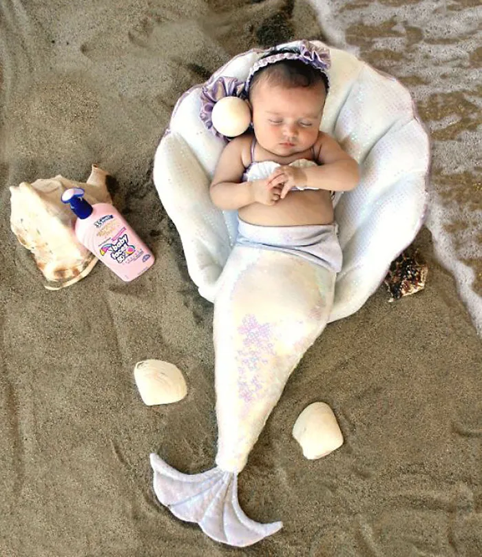 Baby dressed as a mermaid in a shell on the sand, an adorable children's Halloween costume idea for toddlers.