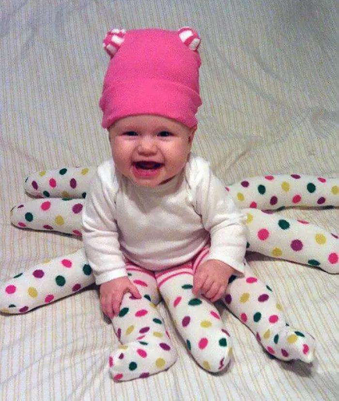 Smiling baby dressed in a creative children's Halloween costume with colorful polka-dot spider legs and a pink hat.