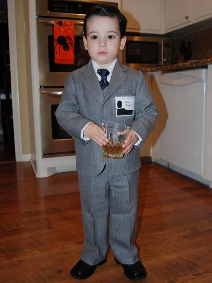 Young boy dressed in a children’s Halloween costume holding a glass, standing in a kitchen setting.