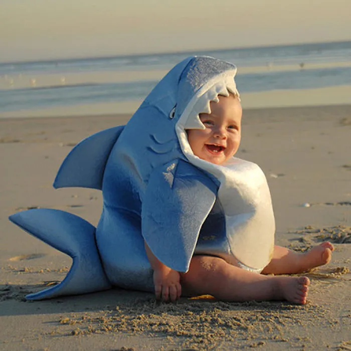 Baby sitting on the beach wearing a cute shark costume, one of the best children’s Halloween costume ideas.