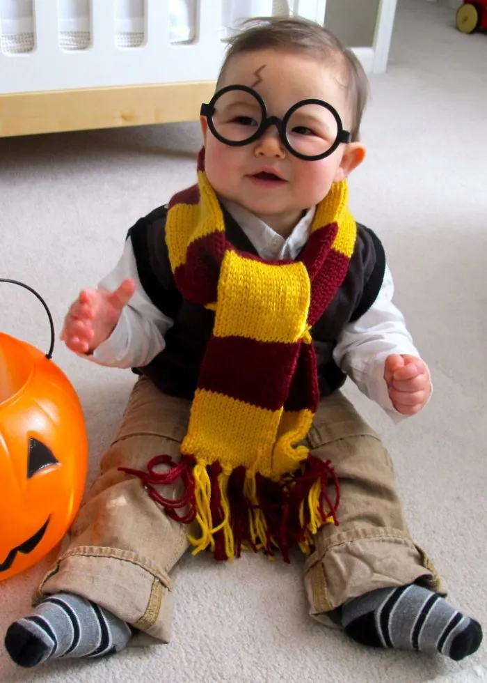 Baby dressed in a children's Halloween costume wearing glasses and a scarf, sitting next to a pumpkin bucket indoors.
