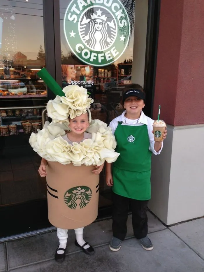 Two children in creative Halloween costume ideas, one dressed as a Starbucks drink and the other as a barista outside a coffee shop.