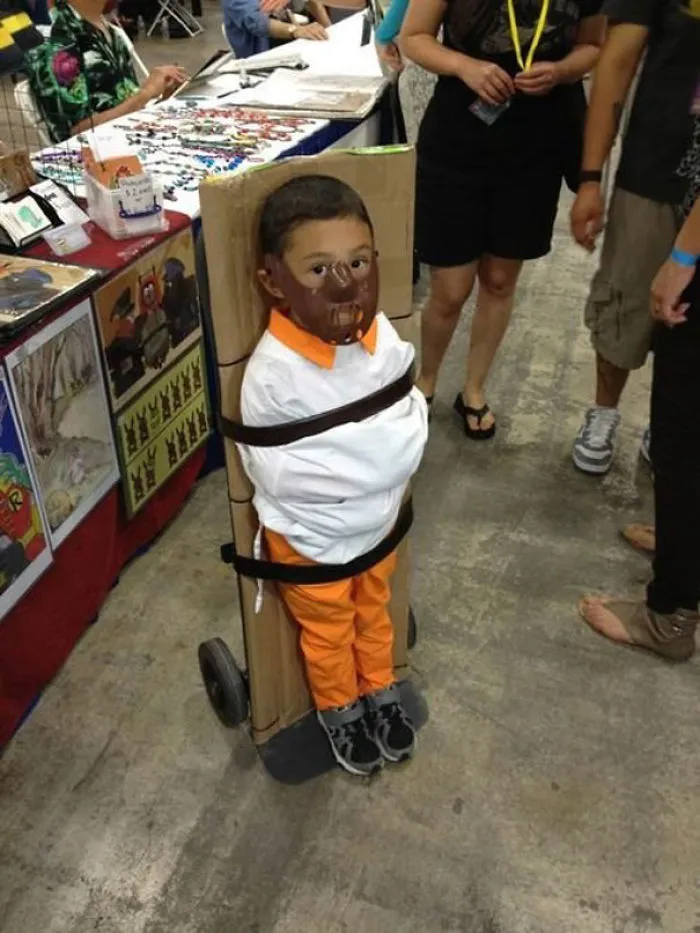 Child in creative Halloween costume wearing orange outfit and face mask, standing in a busy event setting with adults nearby.
