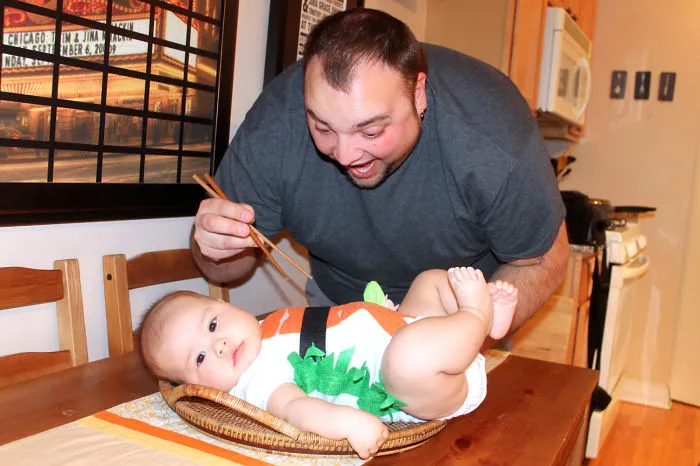 Man playfully using chopsticks near baby dressed in a creative children's Halloween costume idea on a table.
