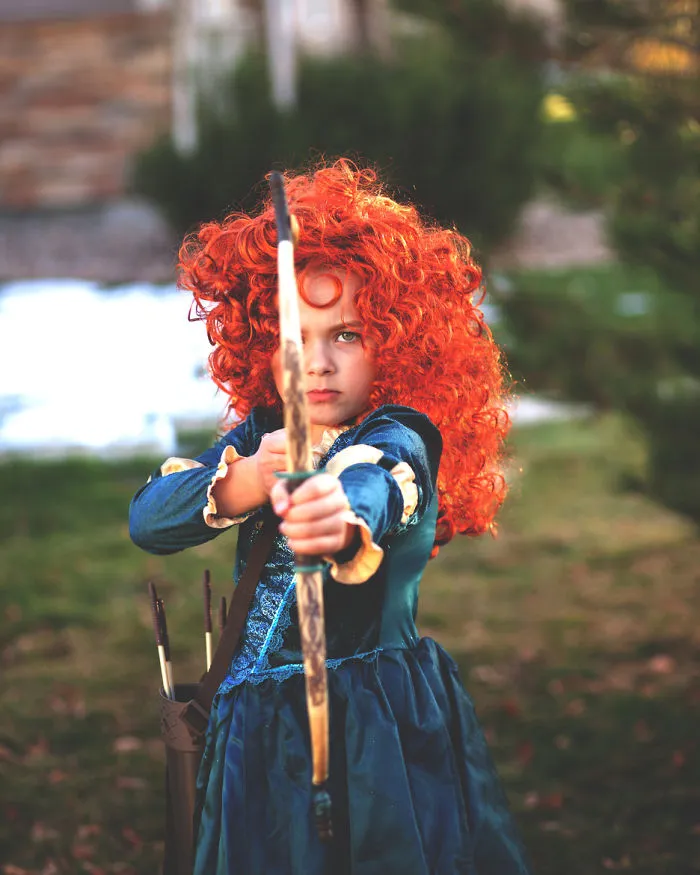 Child in children's Halloween costume ideas with bright red curly hair aiming a bow and arrow outdoors.