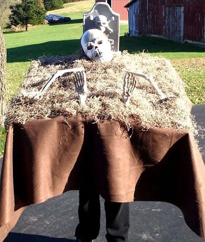 Child's Halloween costume idea featuring a spooky graveyard tombstone with skeleton hands and skull props.