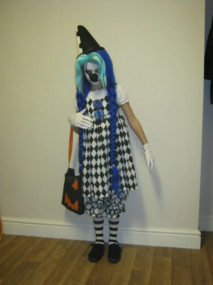 Child in Halloween costume with blue wig, face paint, black and white dress, holding a pumpkin-themed bag for Halloween costume ideas.