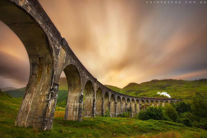 Mystical bridge with arches stretching across green landscape under a dramatic sky, evoking a magical journey to another world.