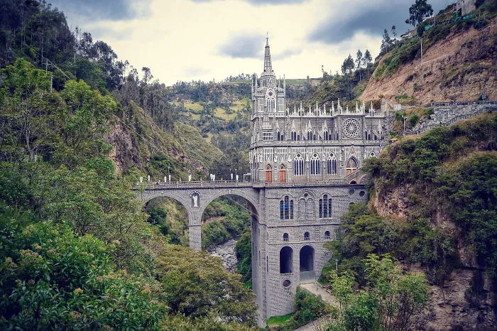 Mystical stone bridge connected to an ornate gothic church surrounded by lush green hills and cloudy sky.