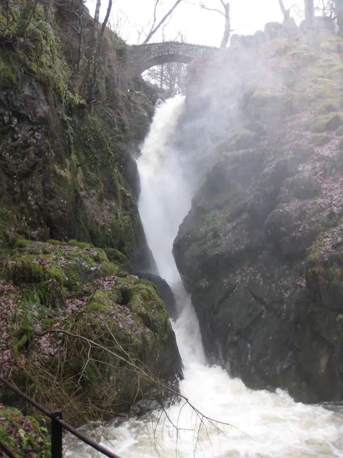 Stone mystical bridge over a powerful waterfall surrounded by moss-covered rocks and mist in a forest setting