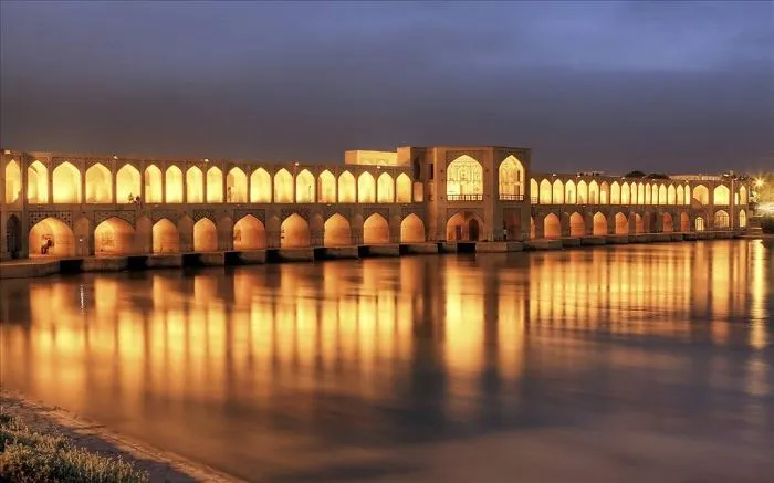 Illuminated mystical bridge over calm water at dusk, reflecting golden lights and creating a magical atmosphere.