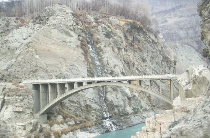 Concrete arch mystical bridge spanning rocky canyon with flowing waterfall and river below in a remote natural landscape