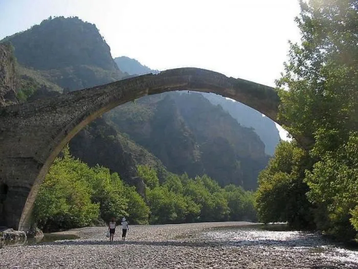Ancient mystical stone bridge arching over rocky riverbed with green trees and mountains in the background.