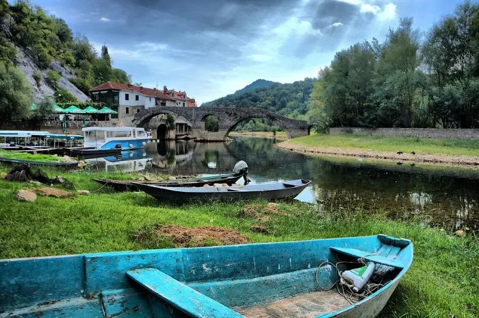 Stone bridge over calm river with boats on grassy shore and forested hills under a cloudy sky, showcasing mystical bridges.