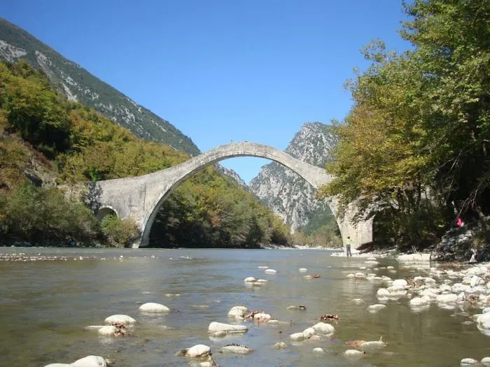 Stone mystical bridge arching over a river surrounded by lush green trees and mountainous landscape under a clear blue sky