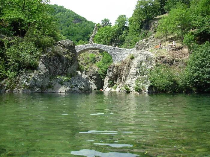 Stone mystical bridge arching over clear green water surrounded by lush trees and rocky cliffs.