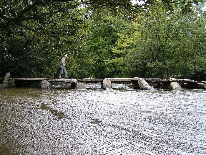Person walking across a mystical bridge made of stone slabs over a flowing river surrounded by lush green trees.