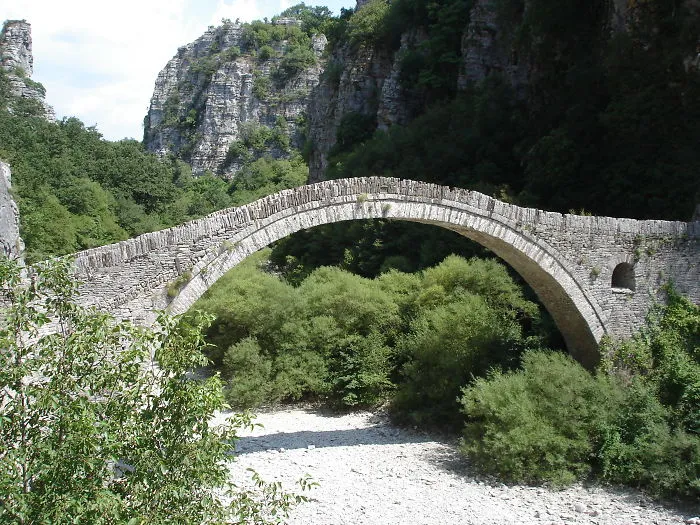 Ancient mystical stone bridge arching over a rocky riverbed surrounded by lush greenery and towering cliffs.