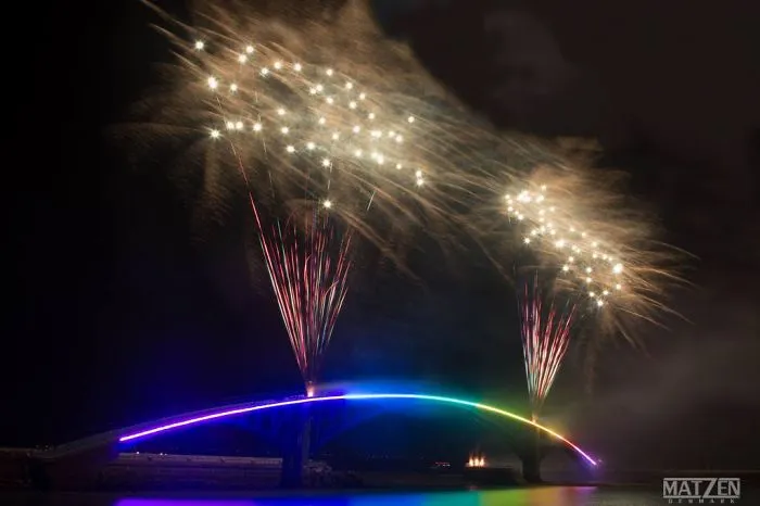 Colorful mystical bridge illuminated at night with fireworks exploding above, creating a magical and otherworldly atmosphere.