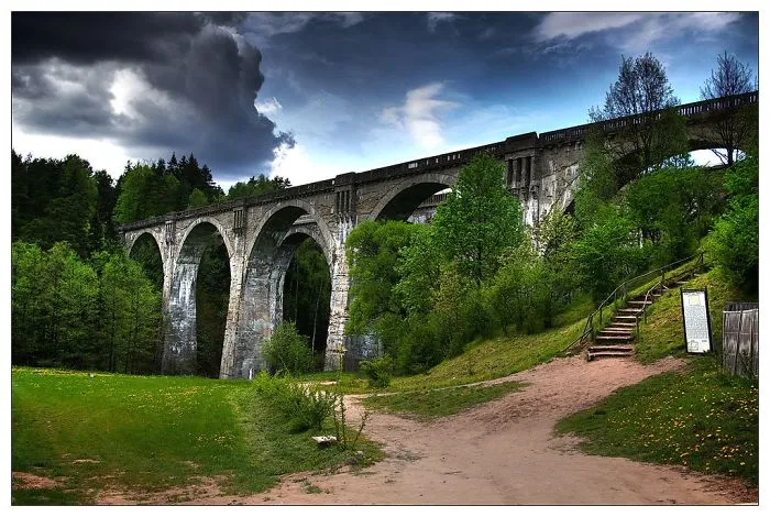 Stone mystical bridge with arches surrounded by lush green trees and a dramatic cloudy sky above.
