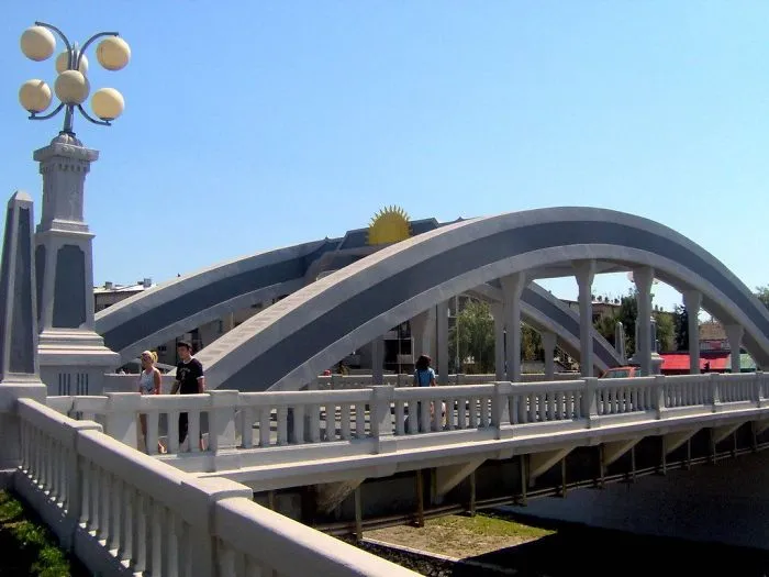 Arched mystical bridge with pedestrians, featuring art deco design and bright lamps under clear blue sky.
