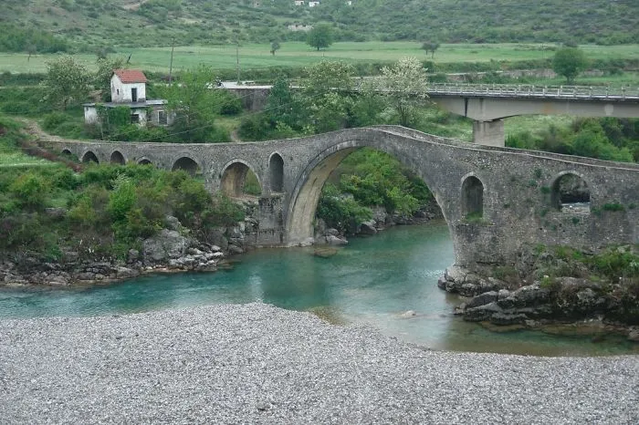 Ancient mystical stone bridge arching over clear river surrounded by lush greenery and rocky riverbank.