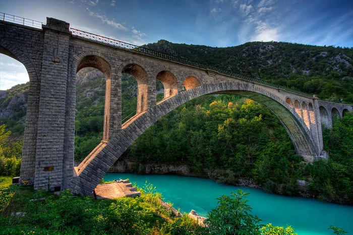 Stone arched mystical bridge over turquoise river surrounded by lush green forest and hills under a blue sky