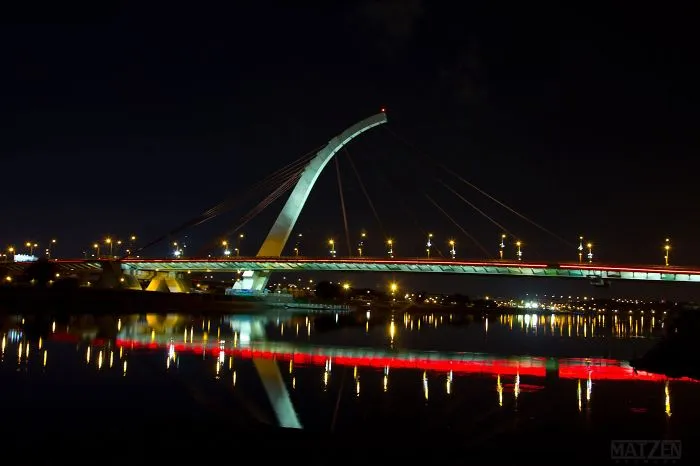 Illuminated mystical bridge at night with colorful lights reflecting on calm water in a serene setting.