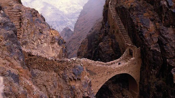 Ancient stone mystical bridge arching over a deep rocky canyon, surrounded by rugged mountainous terrain.