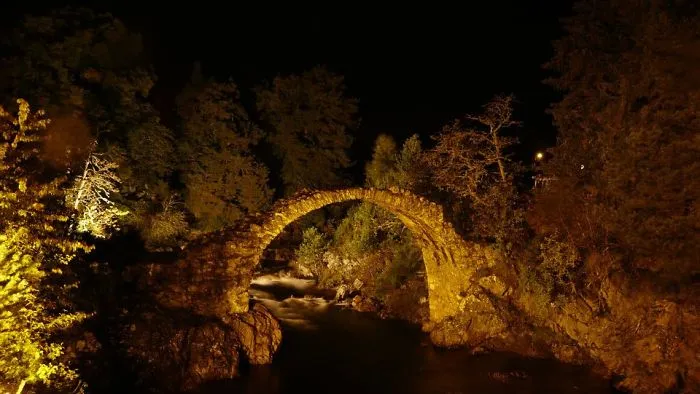 Mystical stone bridge illuminated at night surrounded by trees and flowing river in a magical forest setting.