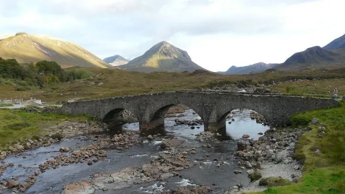 Old stone mystical bridge over flowing river in a lush valley with mountains in the background under a cloudy sky