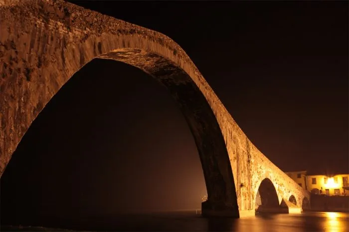 Ancient mystical bridge illuminated at night over calm water, creating a magical and otherworldly atmosphere.