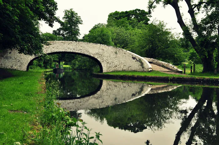 Stone mystical bridge over calm water canal surrounded by lush green trees and grass reflecting in water
