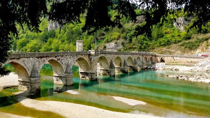 Ancient stone mystical bridge with multiple arches over clear turquoise river surrounded by lush green forest and rocky hills.