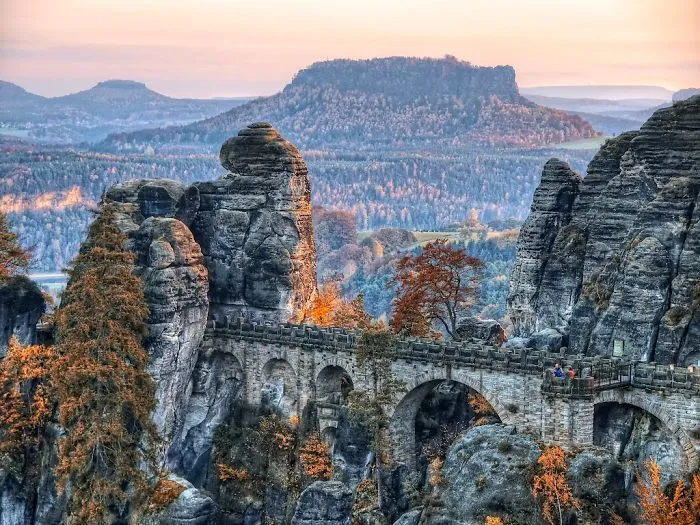 Stone mystical bridge winding through rocky cliffs with autumn trees and misty mountains in the background at sunset.