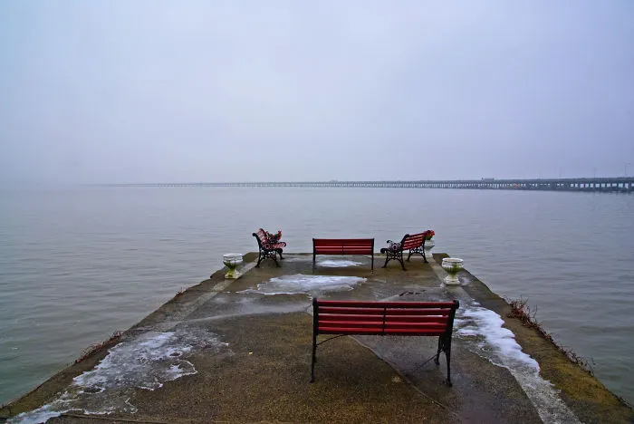 Empty red benches on a misty pier near a long mystical bridge fading into the fog over calm water.