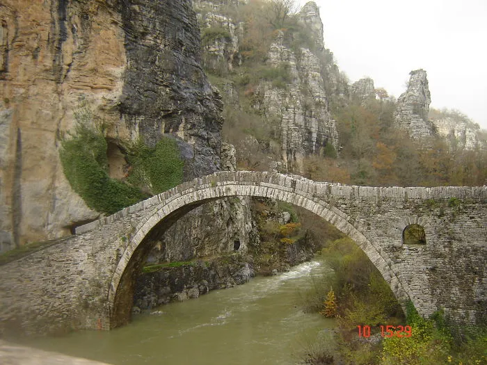 Ancient mystical stone bridge arching over a river, surrounded by rugged cliffs and autumn foliage in a remote landscape