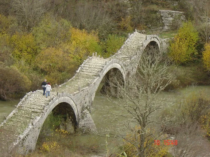Ancient mystical stone bridge with multiple arches surrounded by autumn trees and flowing river below, two people walking across.