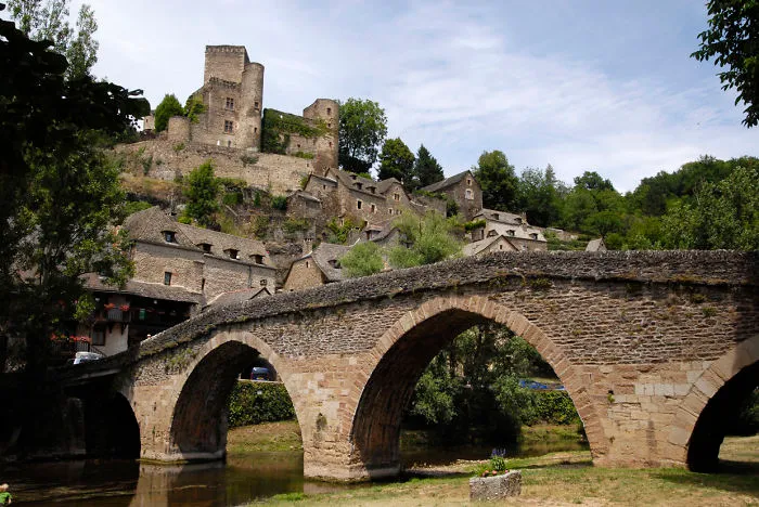 Ancient mystical stone bridge with arches over river, leading to old village and castle surrounded by greenery.