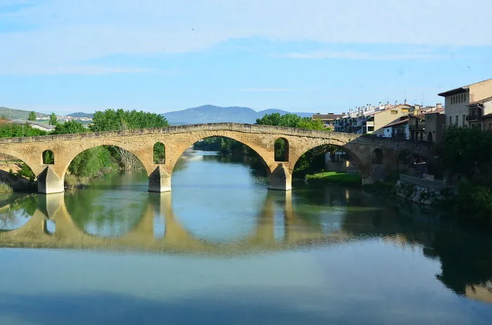 Ancient mystical stone bridge with multiple arches spanning a calm river under blue sky and distant hills.