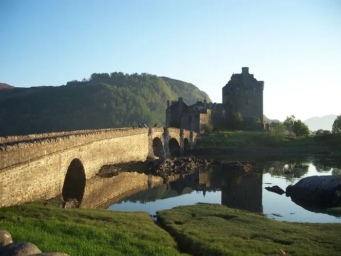 Stone bridge over calm water leading to a historic castle, surrounded by lush green hills under a clear sky.
