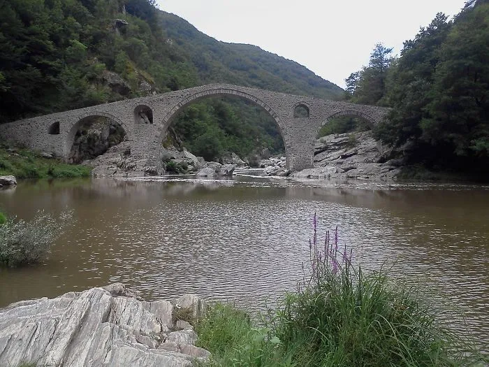 Ancient mystical stone bridge arching over a river surrounded by lush green trees and rocky landscape.