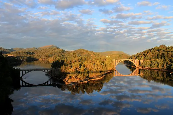 Two mystical bridges arching over calm water surrounded by forested hills under a partly cloudy sky.