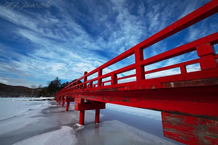 Red mystical bridge extending over a frozen lake under a vast blue sky with wispy clouds.